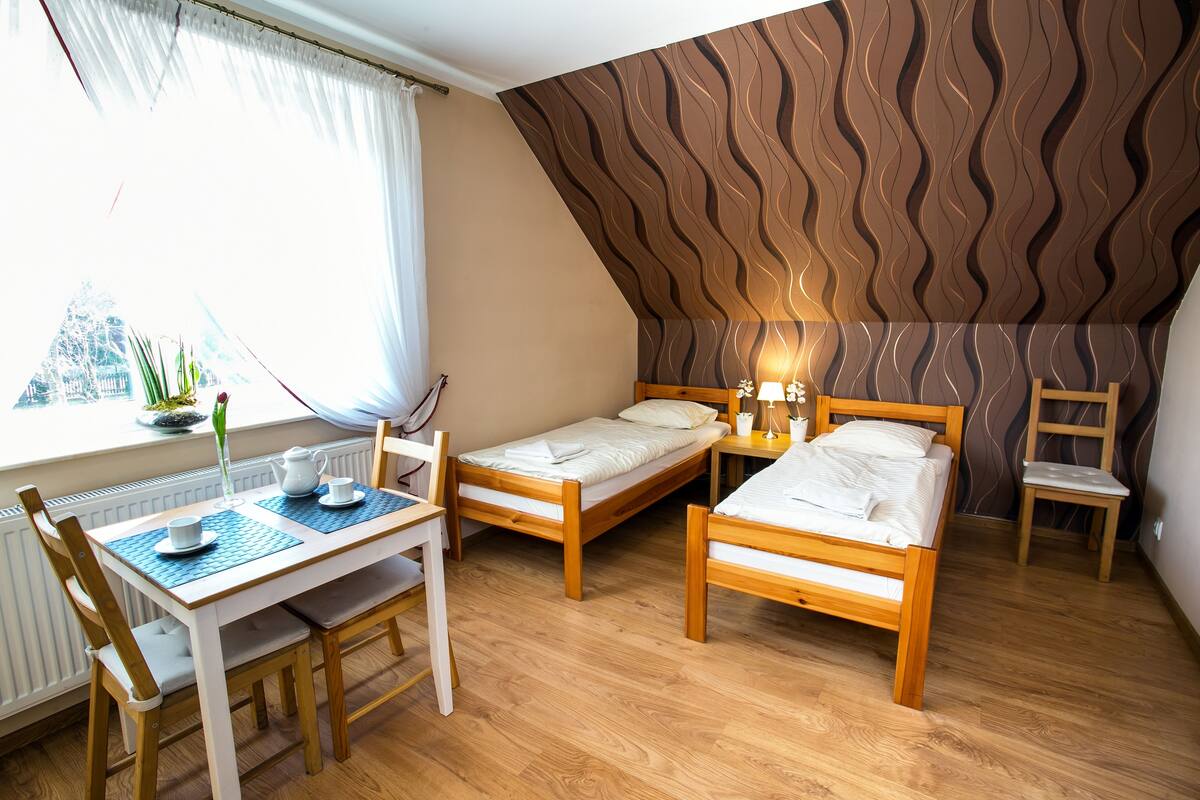 A small, cozy room with two wooden beds, a table with chairs, and a window with curtains.
