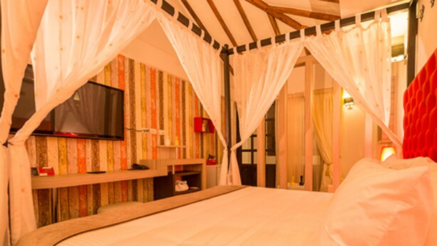 A bedroom with a canopy bed, a television on the wall, and a red accent wall. - Hotel Capitán Ricaurte (Villa de Leyva)