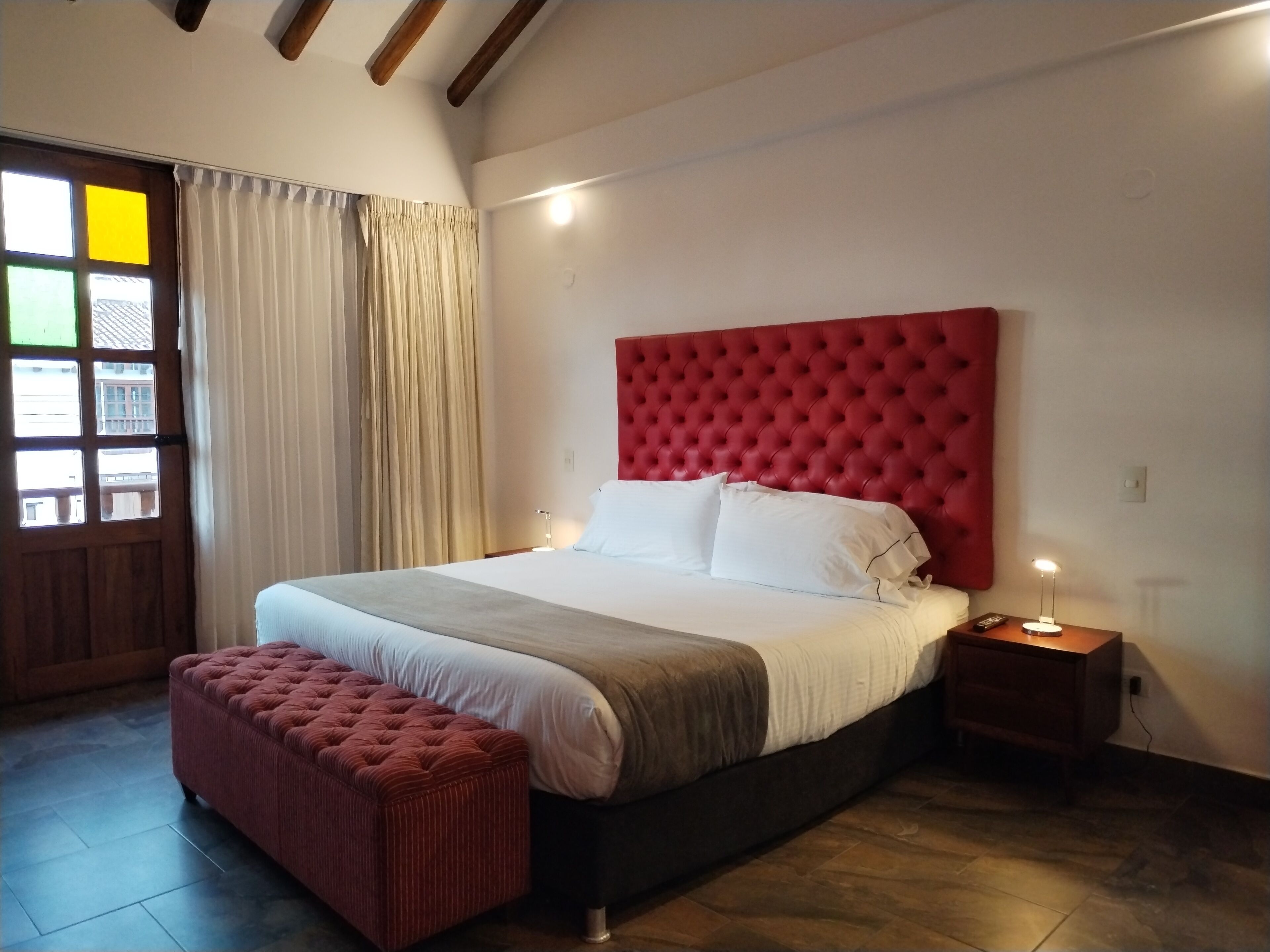 A bedroom with a large bed, a red upholstered headboard, a bedside table with a lamp, and a red bench.