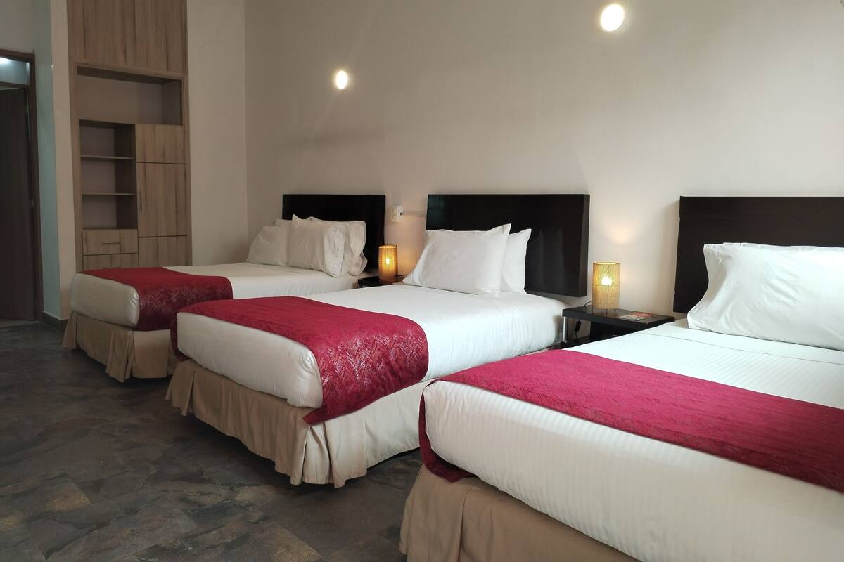 Habitación de hotel con tres camas, cada una con ropa blanca y cojines rojos. Se observan armarios de madera y una mesita con una lámpara.
