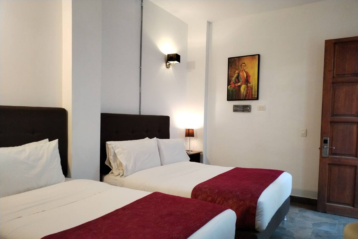 Habitación de hotel con dos camas, una mesita de noche con una lámpara, un cuadro en la pared y una puerta de madera.