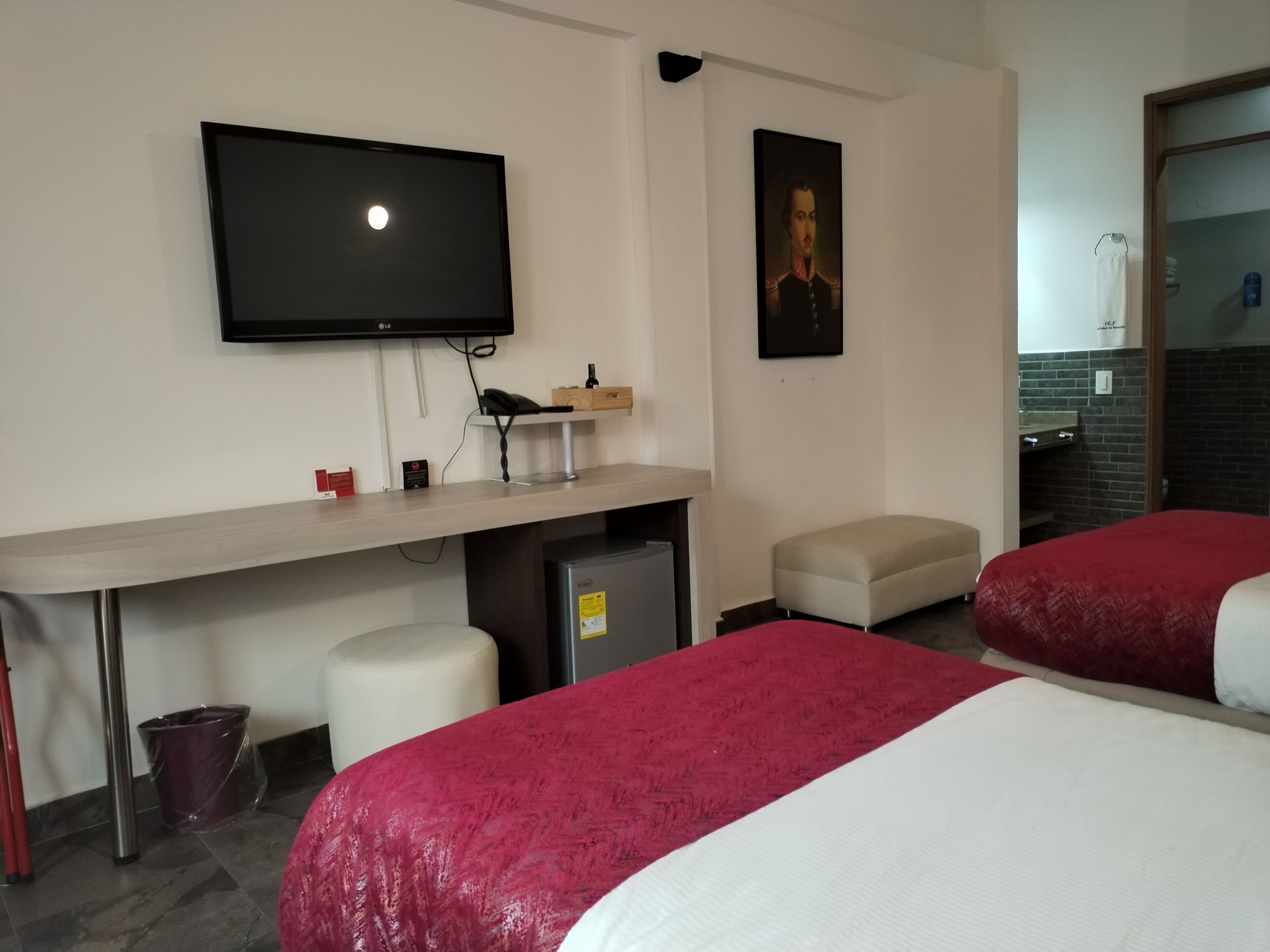 Una camera d'albergo con un letto, una scrivania con televisore, un piccolo frigorifero, un cestino e un quadro appeso al muro.