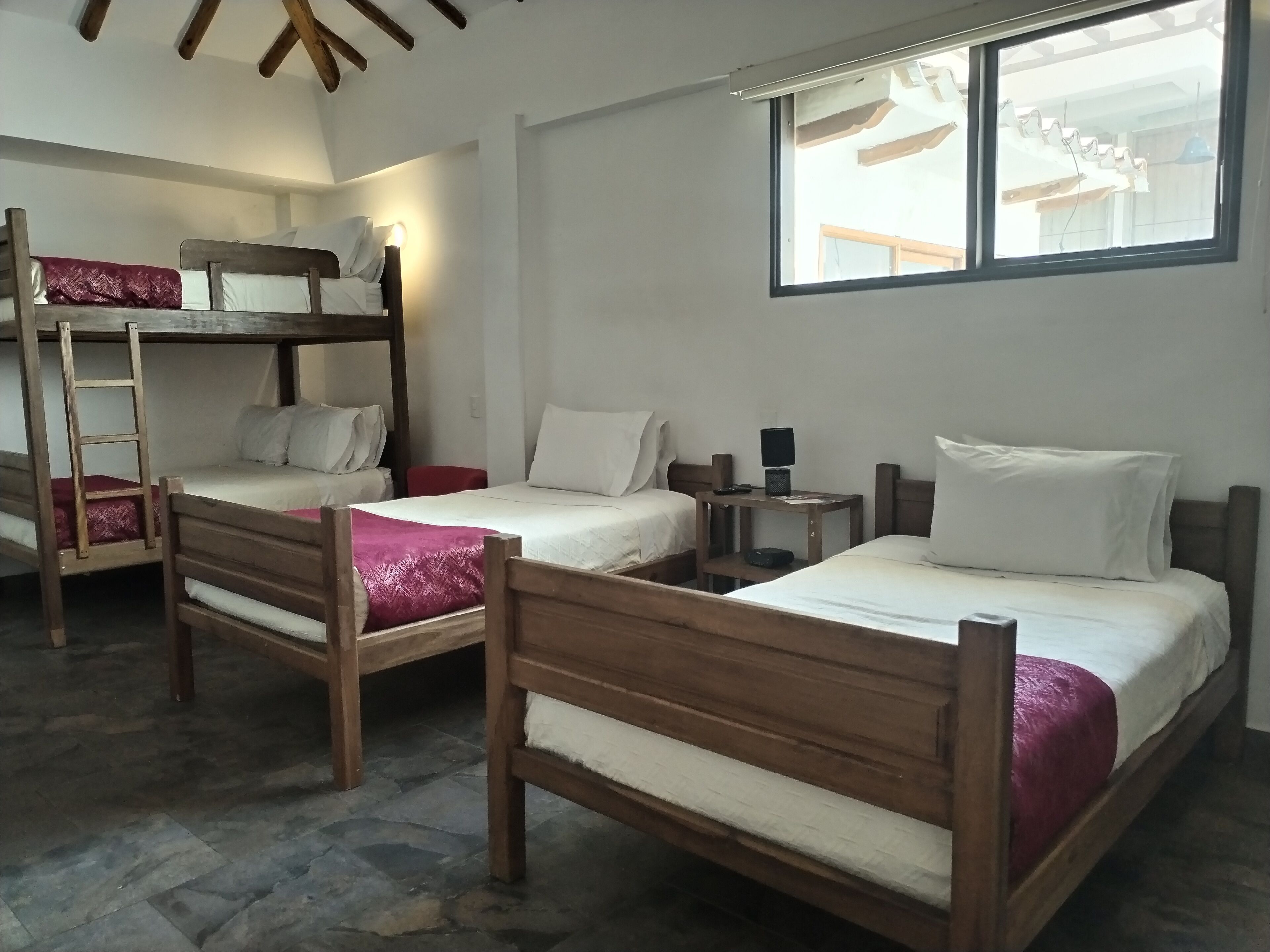A dormitory room with bunk beds, a small table with a lamp, and a window with a view of the outside.