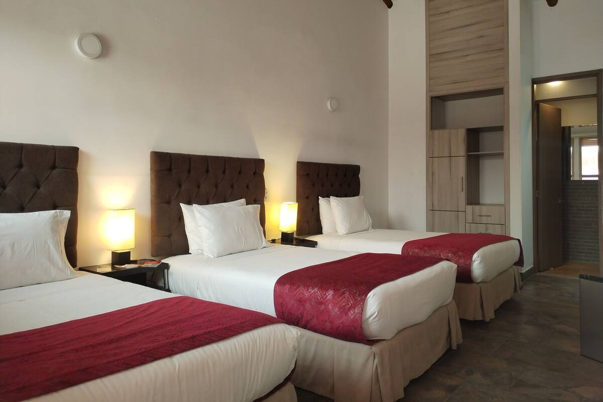 Habitación de hotel con tres camas, cada una con una manta roja, ropa de cama blanca y un cabecero marrón. Cada cama tiene una mesita de noche con una lámpara, y se aprecia un armario de madera al fondo.