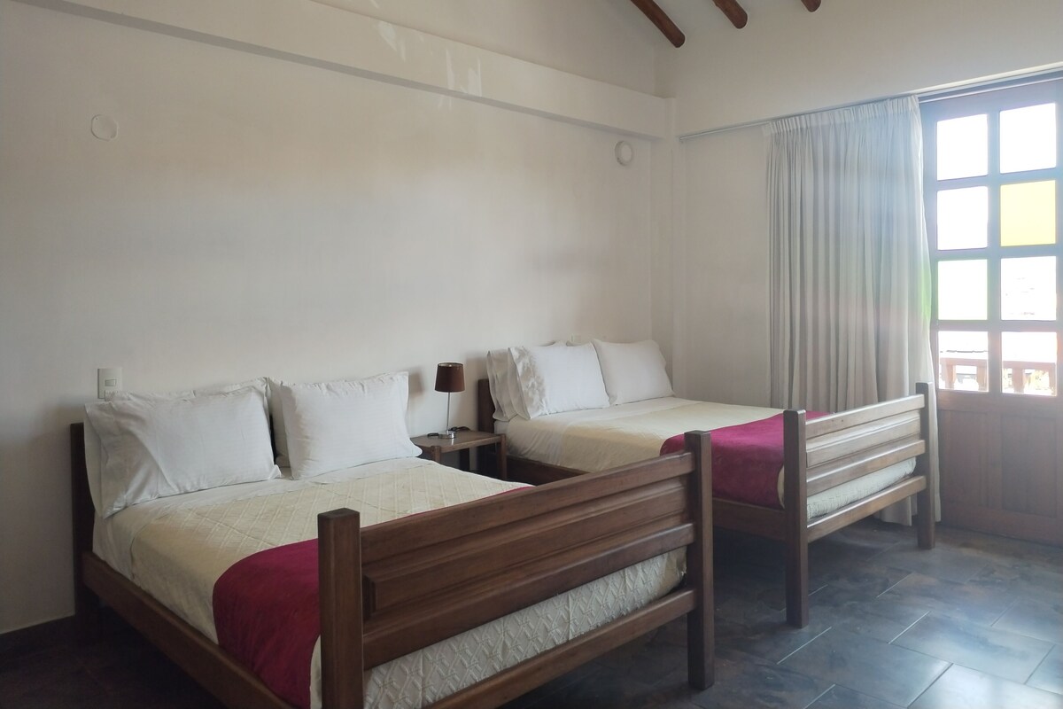 A room with two wooden beds, white bedding, a nightstand with a lamp, and a window with curtains.