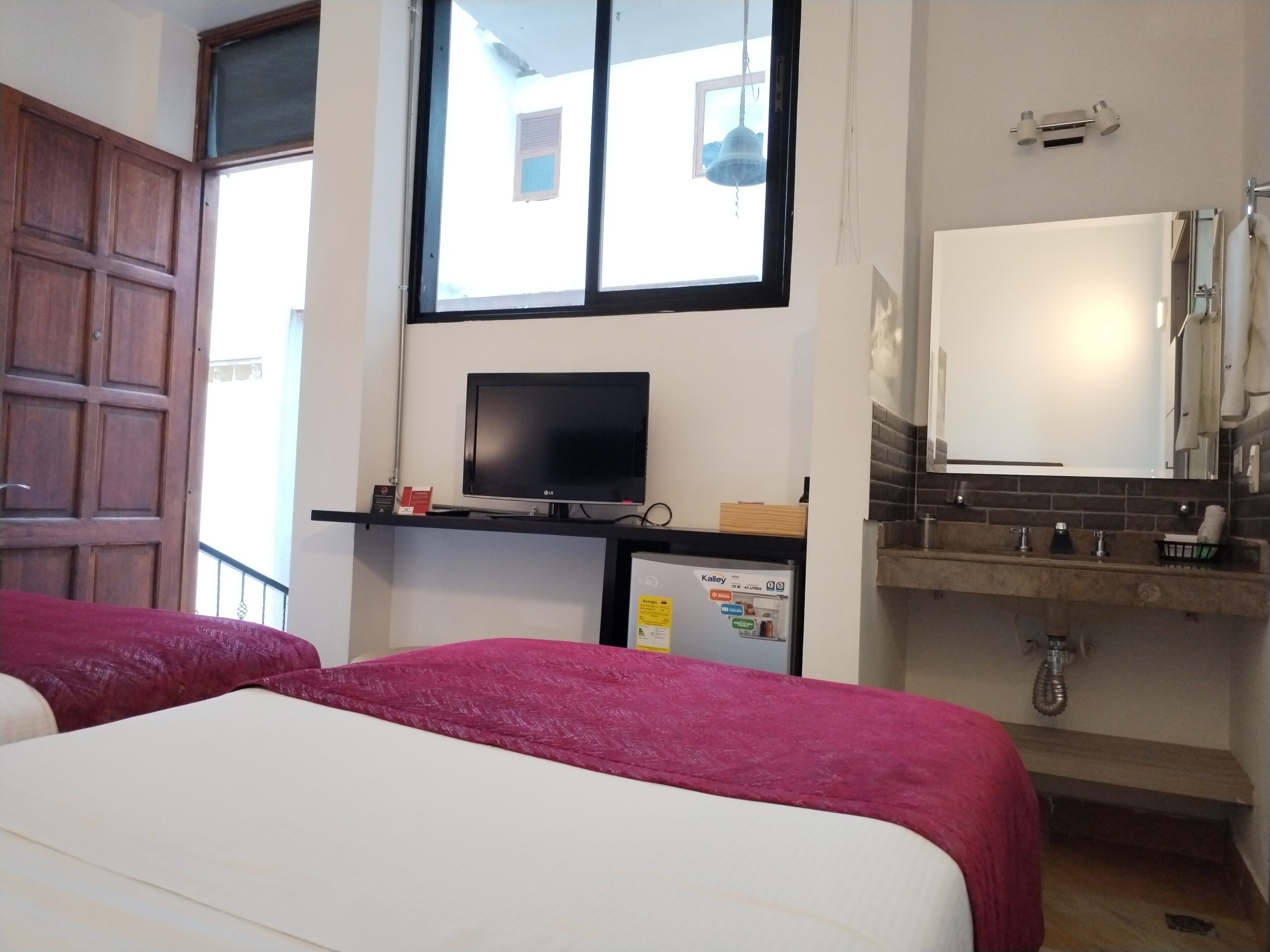 A hotel room with two beds, a television, a small refrigerator, a bathroom with a sink and mirror, and a window with a view of buildings outside.