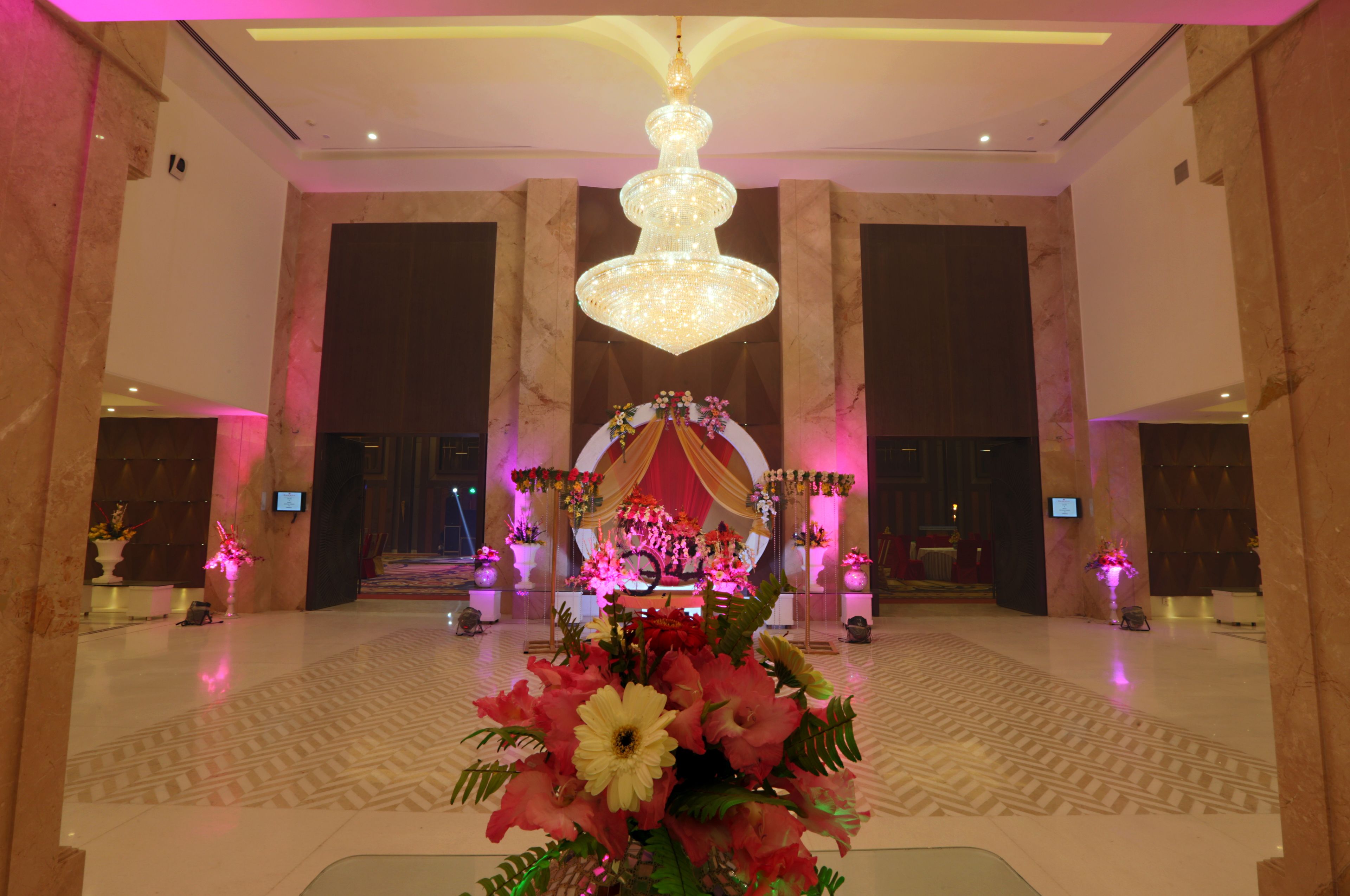 A spacious lobby with a grand chandelier, a stage with floral decorations, and a patterned floor.