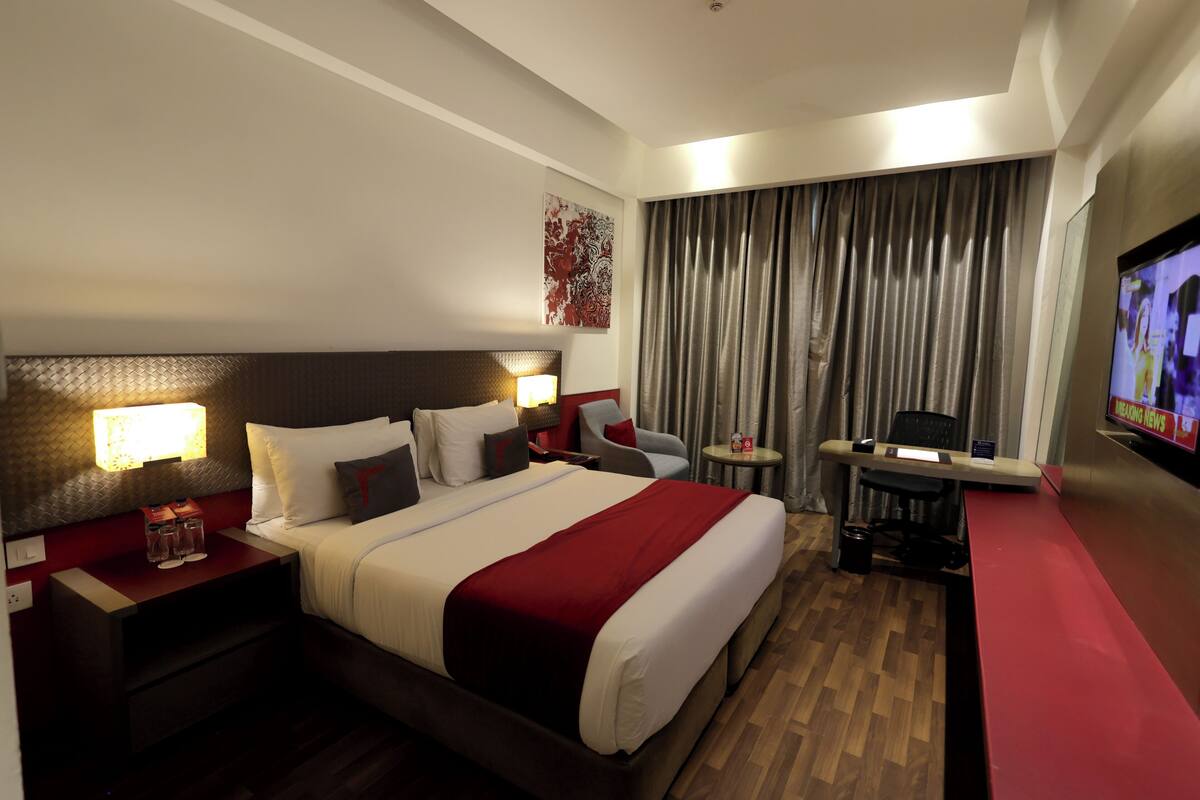 A hotel room with a large bed, bedside tables with lamps, a desk with a chair, a TV, and a window with curtains.