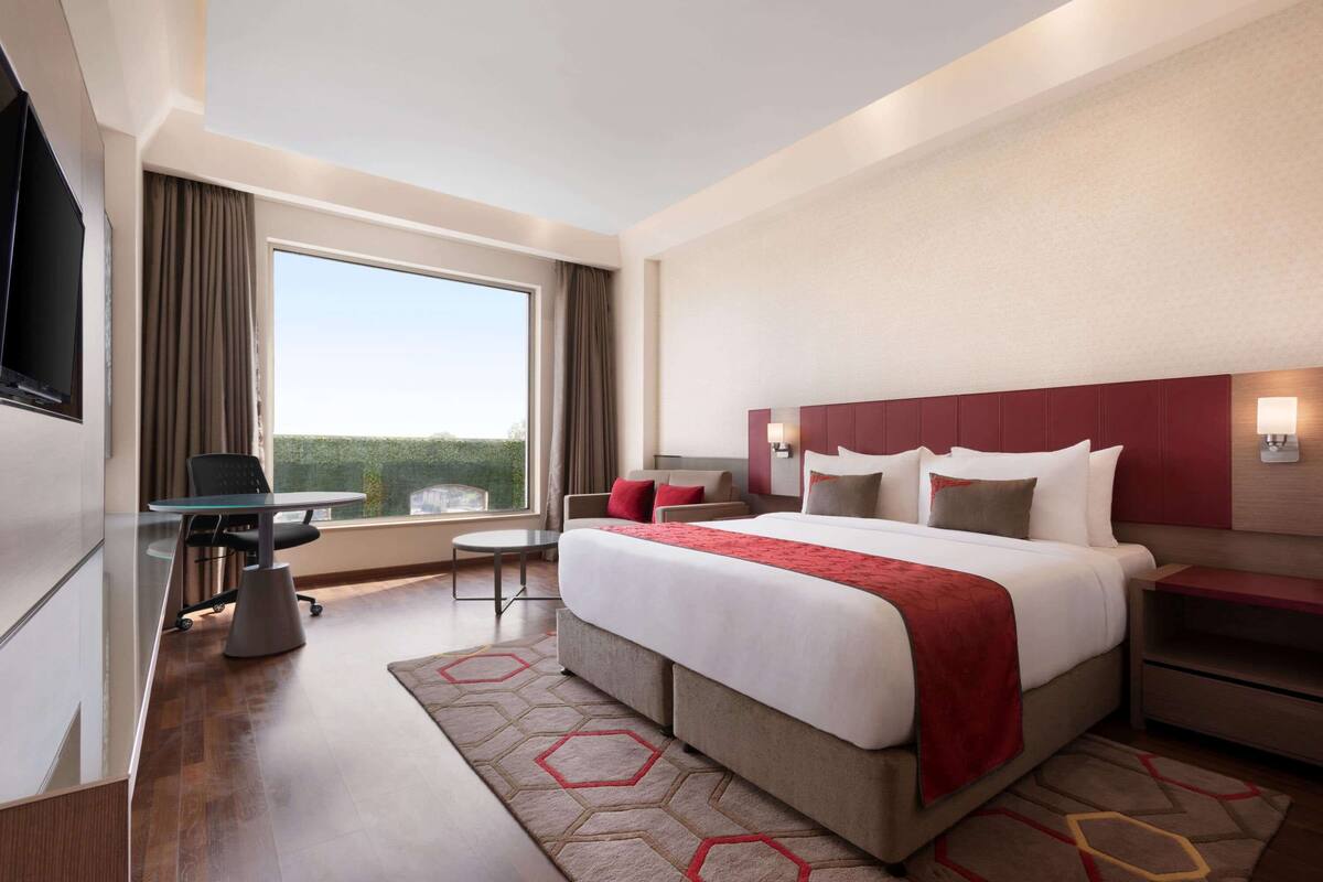 A hotel room with a large bed, a desk with a chair, a small table, and a view of greenery through the window.