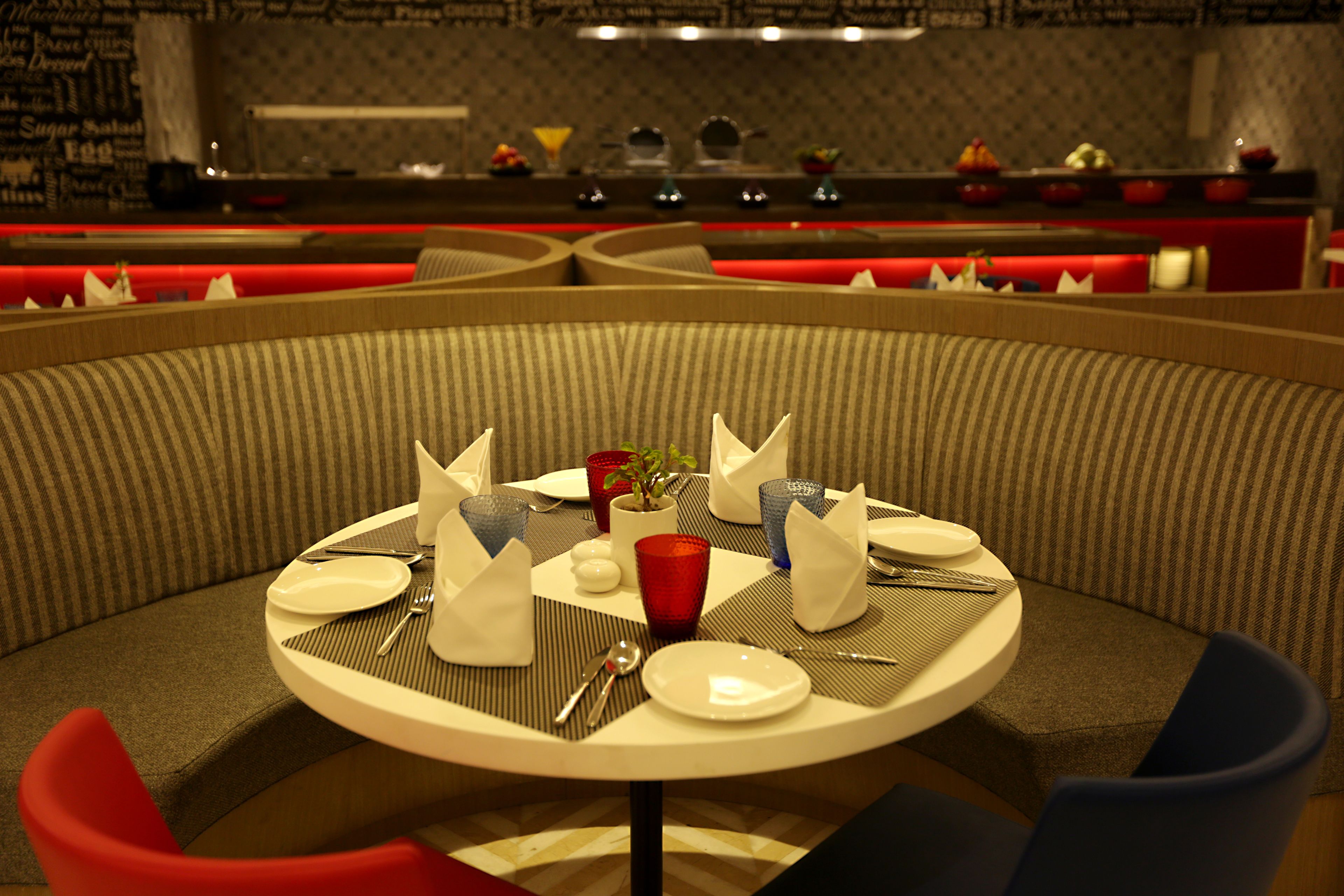 Comedor con una mesa redonda dispuesta para dos personas, adornada con un florero y planta, copas rojas y azules, y cubiertos ordenados.