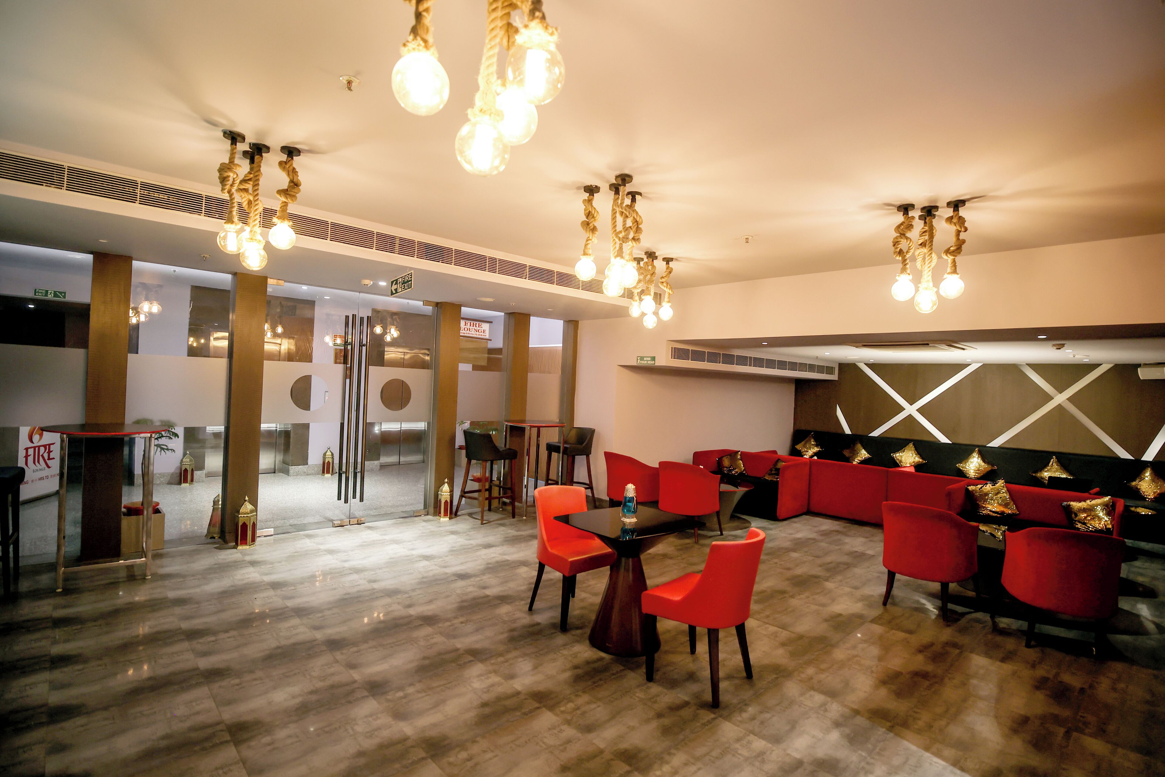A spacious lobby with a modern design, featuring a central seating area with red chairs and a black table, and a decorative ceiling with hanging light fixtures.