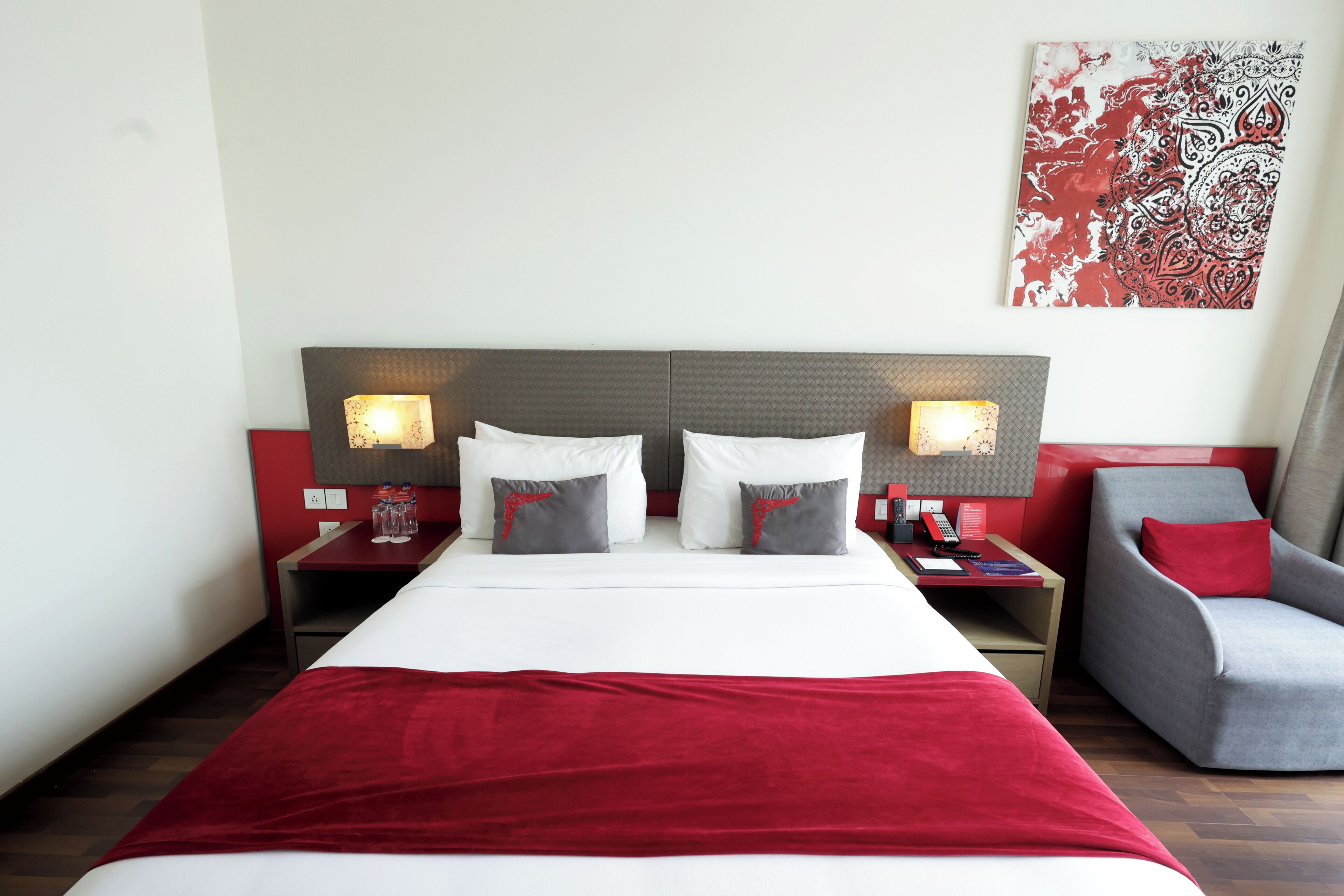 Habitación de hotel con una cama grande, dos mesitas de noche con lámparas, una silla y una obra de arte con motivos rojos y blancos en la pared.