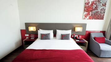 Habitación de hotel con una cama grande, dos mesitas de noche con lámparas, una silla y una obra de arte con motivos rojos y blancos en la pared.
