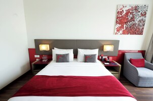Habitación de hotel con una cama grande, dos mesitas de noche con lámparas, una silla y una obra de arte con motivos rojos y blancos en la pared.