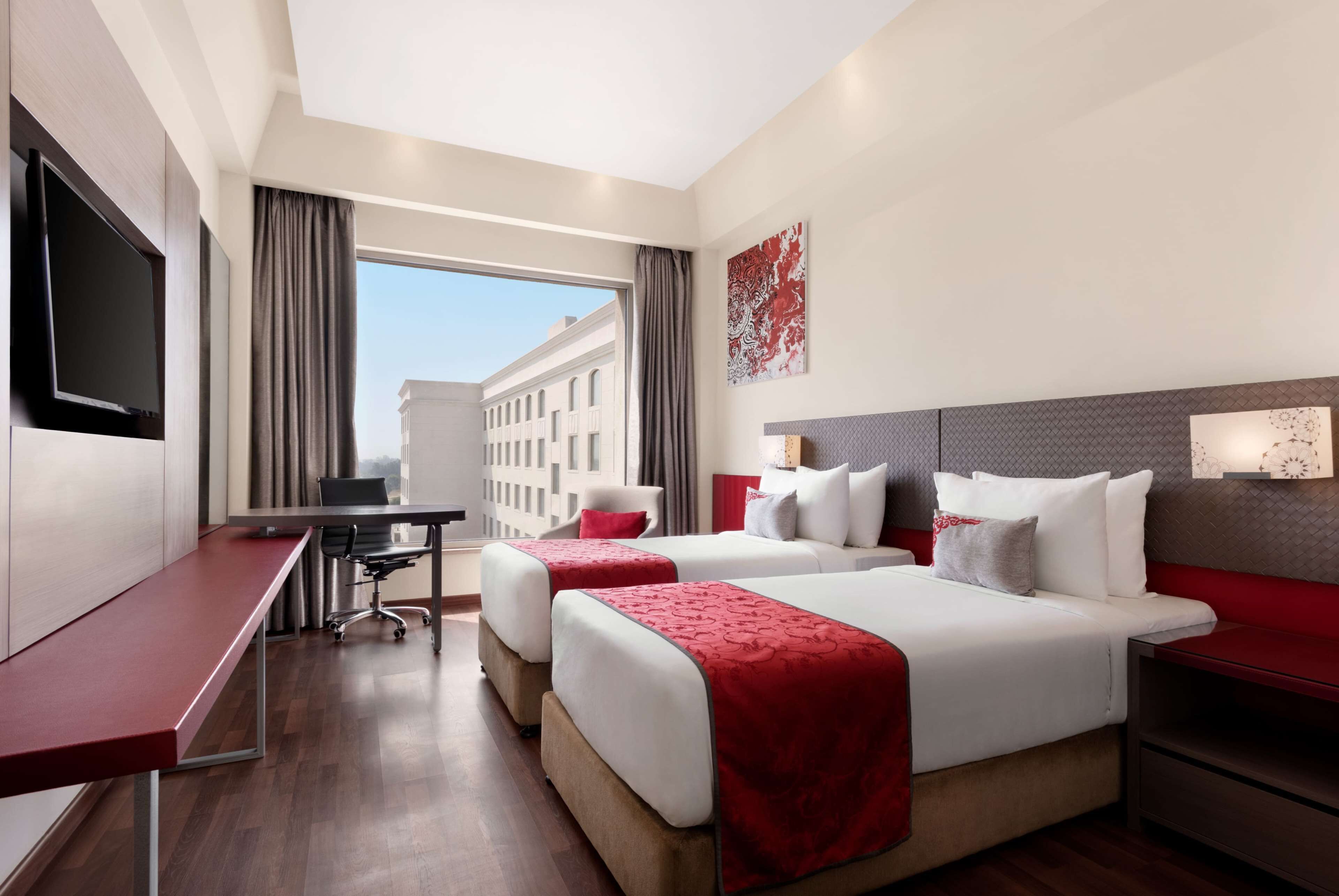 A hotel room with two beds, a desk, a TV, and a large window offering a city view.