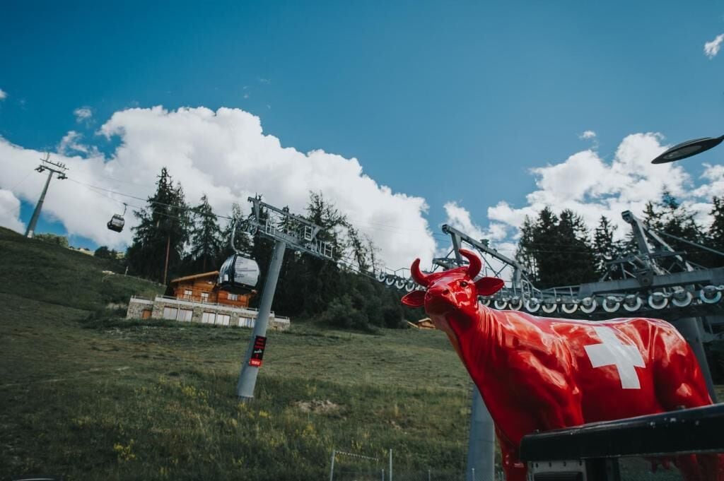 Een rode stierstandbeeld met een wit kruis op de rug, een stoeltjeslift en een klein gebouw met een rood dak op de achtergrond.