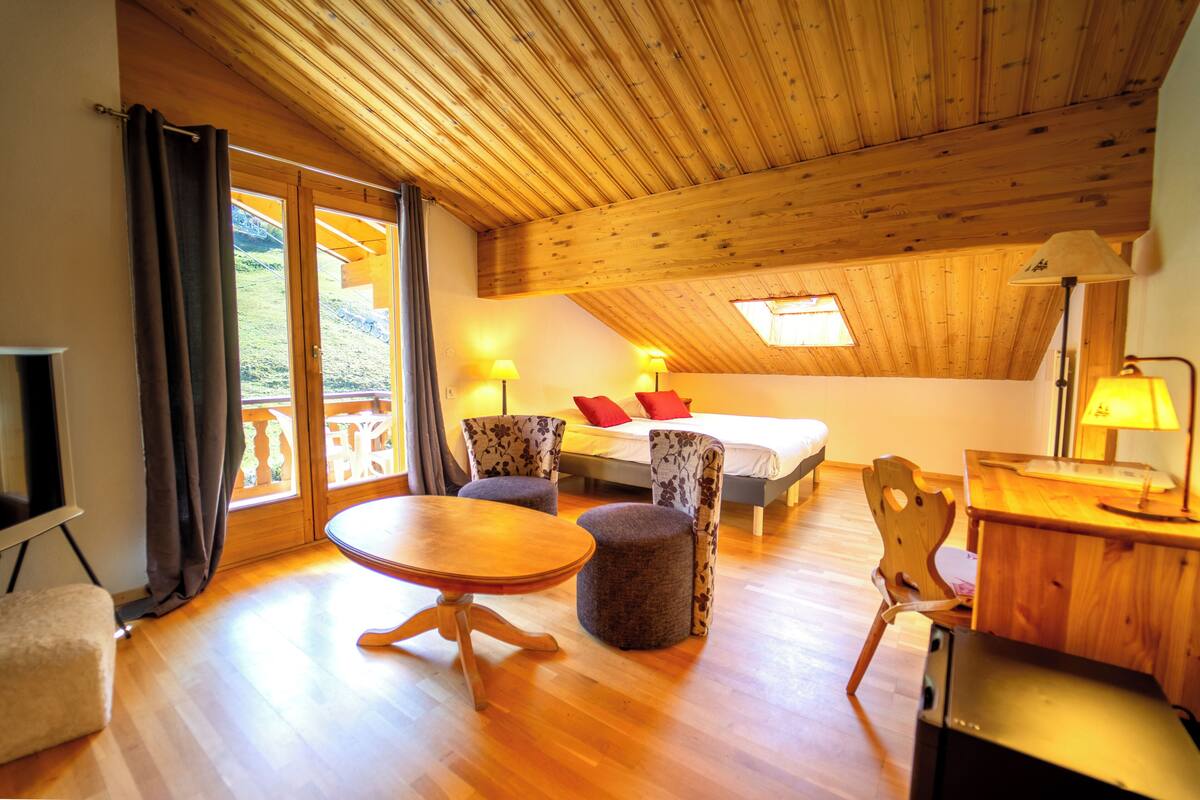 A cozy room with a wooden ceiling, a bed with red pillows, a desk with a lamp, and a view of a balcony.
