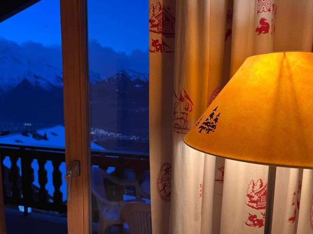 Ein Zimmer mit Blick auf verschneite Berge durch ein Fenster, Vorhänge mit traditionellen Mustern und eine Lampe.