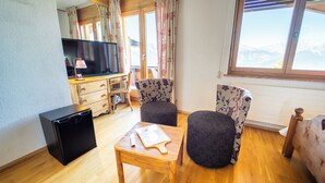 Een compacte kamer met houten vloer, een kleine zwarte koelkast, een houten tafel met een snijplank, twee gestoffeerde stoelen, een televisie en een raam met uitzicht op bergen.
