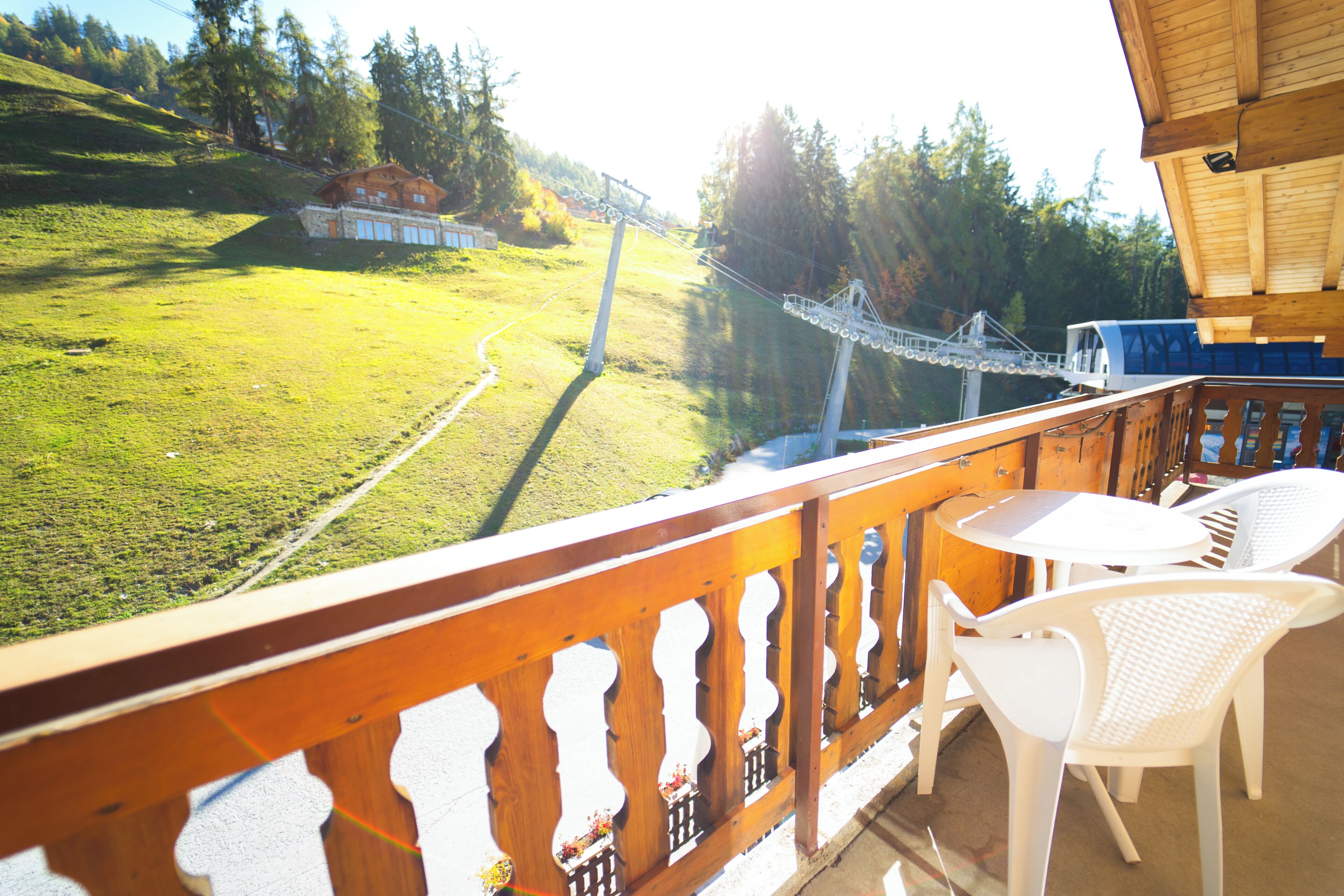 A balcony with a wooden railing, white plastic chairs, and a view of a green landscape with a ski lift.
