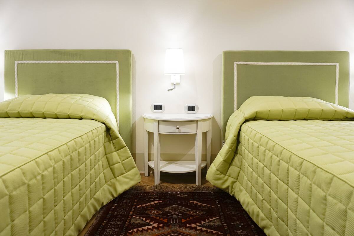Une chambre avec deux lits, chacun recouvert d’une couette matelassée verte et agrémenté d’une tête de lit assortie. Une petite table de chevet blanche avec une lampe se trouve entre les deux lits.