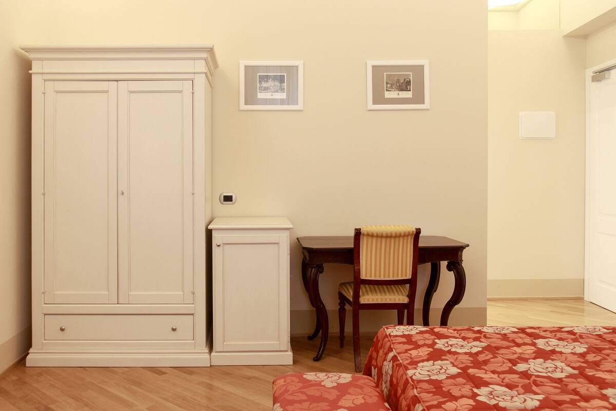 Une chambre avec une grande armoire, un bureau avec une chaise, deux tableaux encadrés au mur et un lit recouvert d’une couvre-lit à motifs floraux.