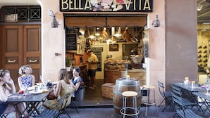 Un área de comedor al aire libre con mesas y sillas, un letrero que dice "Bella Vita" y una persona de pie en la entrada.