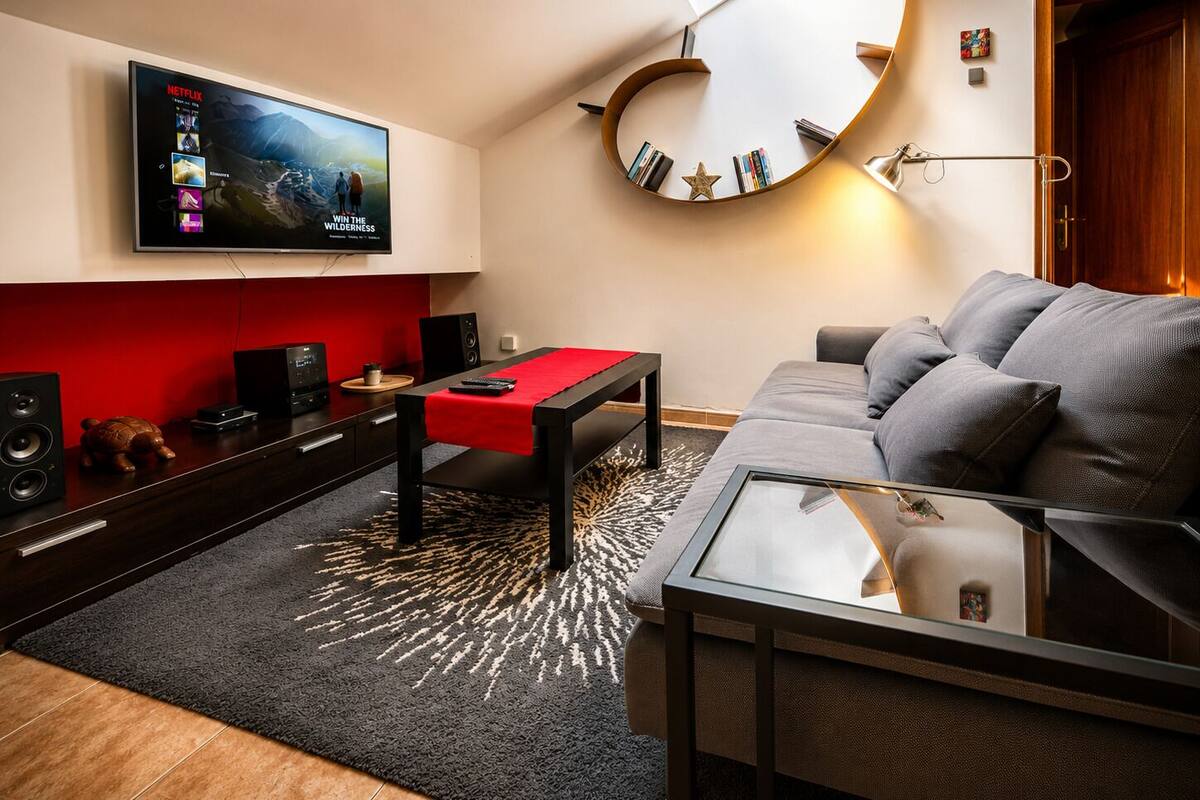 A modern living room with a flat-screen TV, a round mirror, a sofa, a coffee table, and a red accent table.