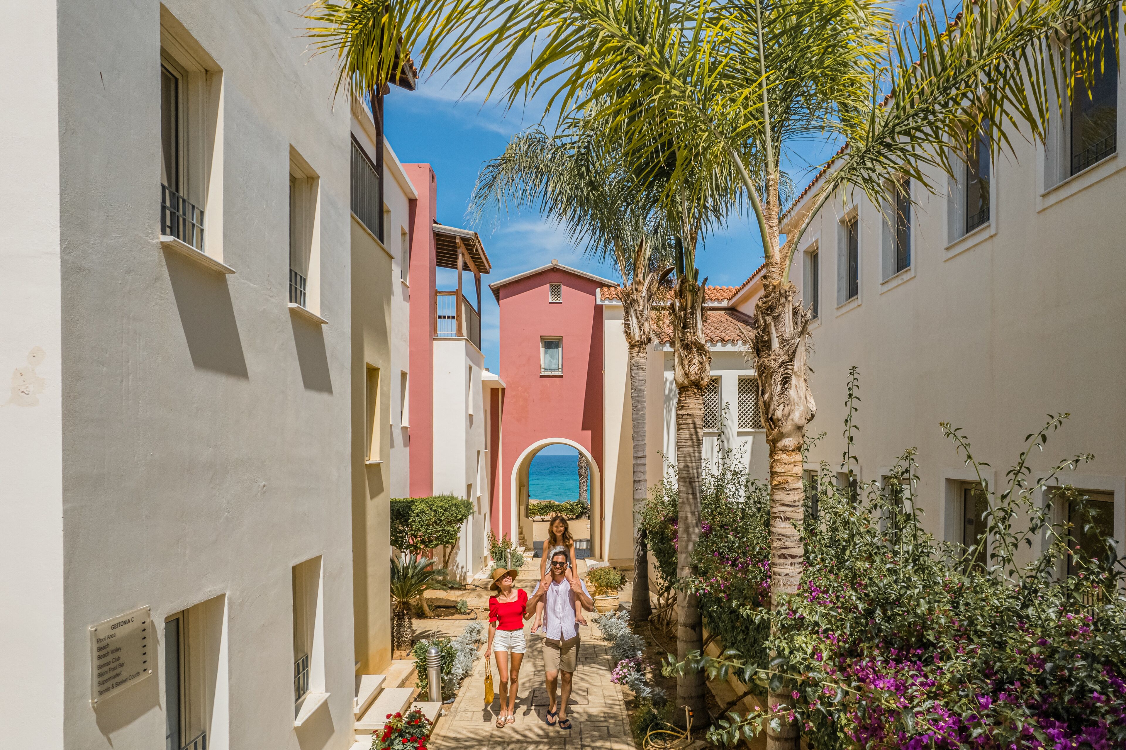 A narrow street with buildings on either side, a palm tree, and a view of the sea through an archway.