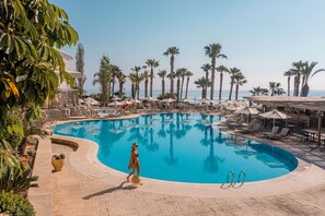 A large, curved swimming pool with lounge chairs and umbrellas, surrounded by palm trees and a clear sky. - Louis Althea Beach Hotel (Paralimni)