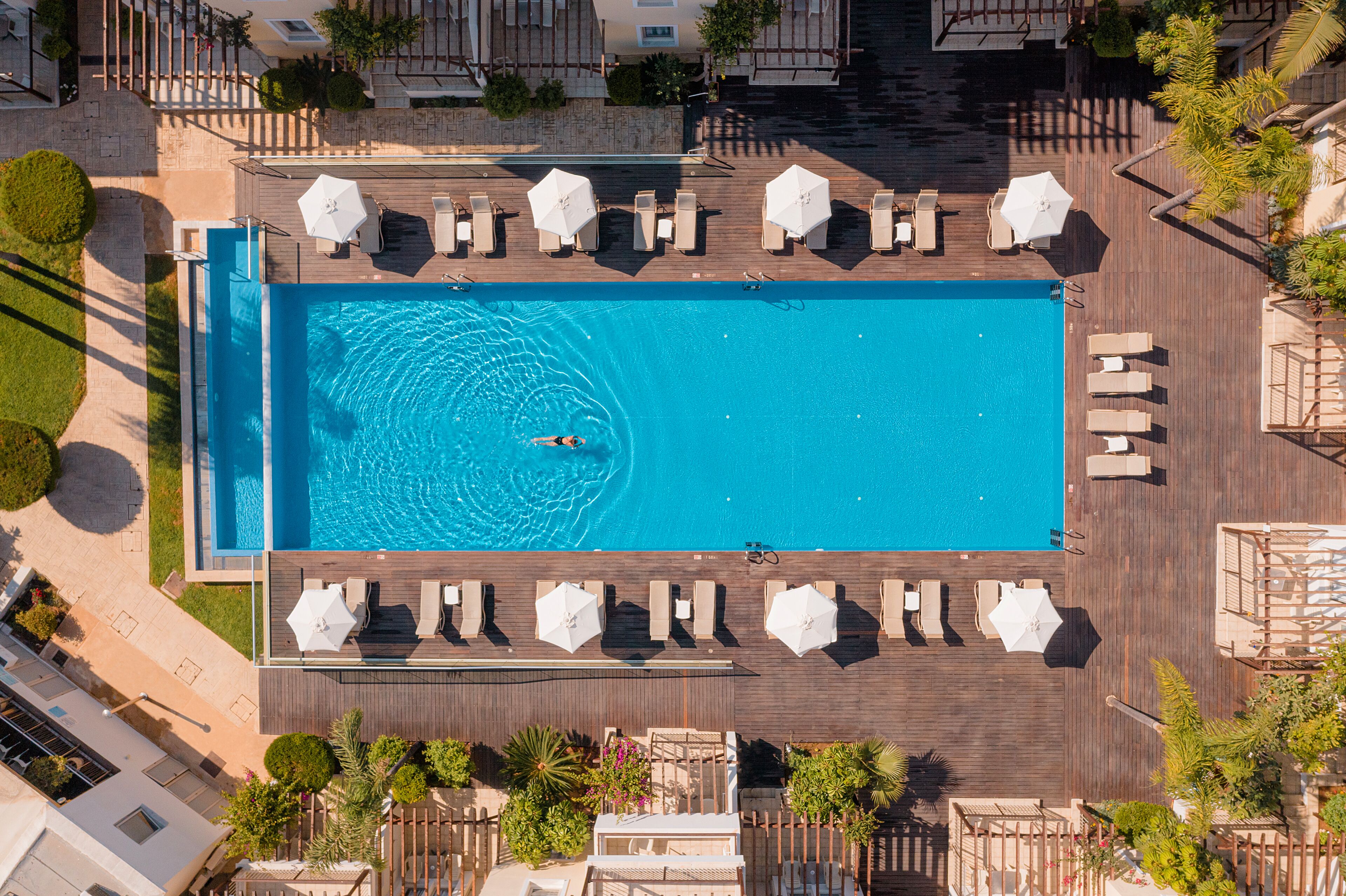 An aerial view of a rectangular swimming pool with a person swimming, surrounded by lounge chairs and umbrellas on a wooden deck.
