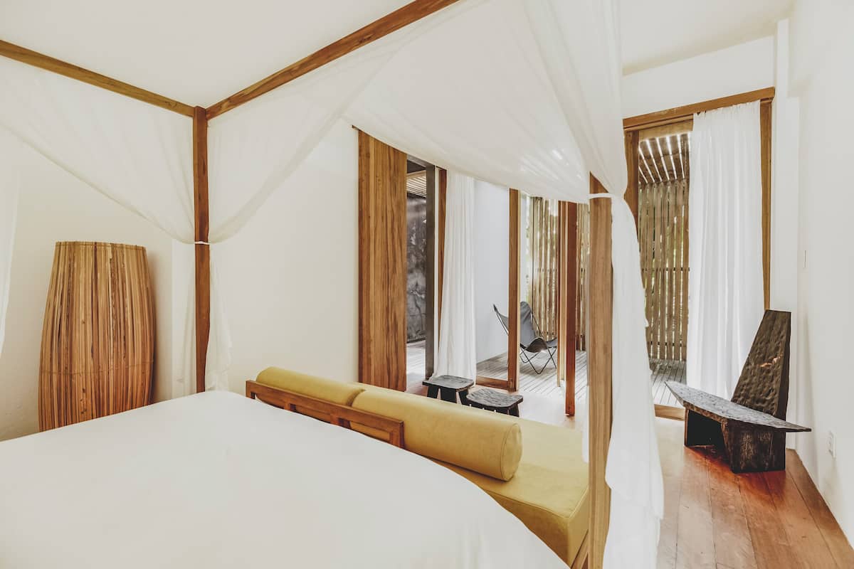 Un dormitorio con una cama grande, un sofá, una mesita y una silla. Hay una ventana con cortinas y vistas al exterior.