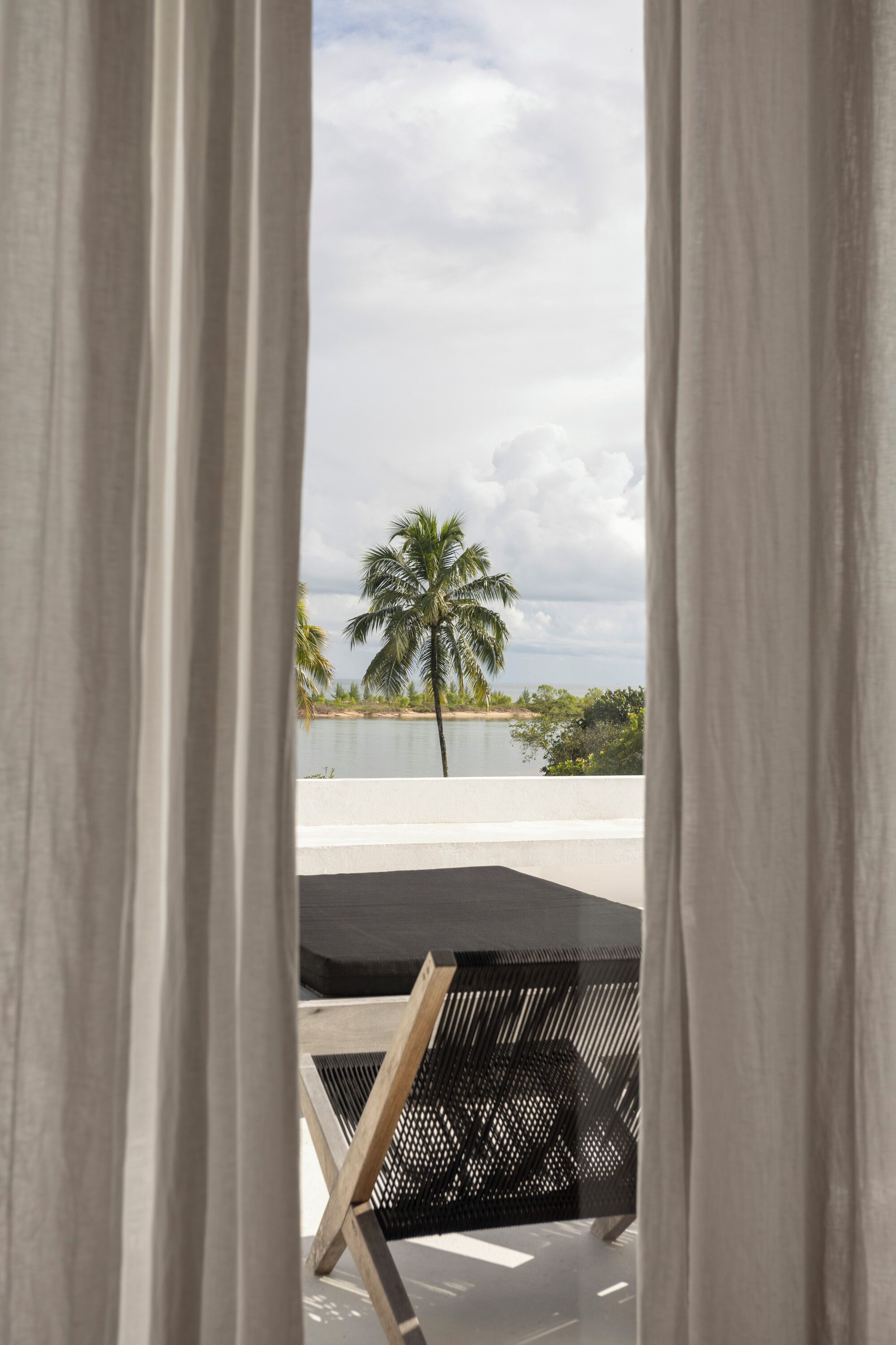A view through curtains of a room with a black outdoor seating area, a palm tree, and a beach in the background.