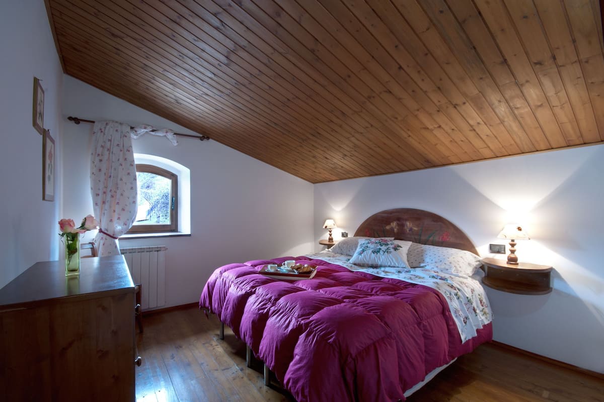 Un dormitorio con techo de madera, una cama con colcha morada, dos lámparas de noche, una ventana con cortina y un aparador con un jarrón de flores.