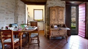 A rustic dining area with a wooden table, chairs, and a cabinet. A kitchen is visible in the background. - Fonte al Noce (Gubbio)
