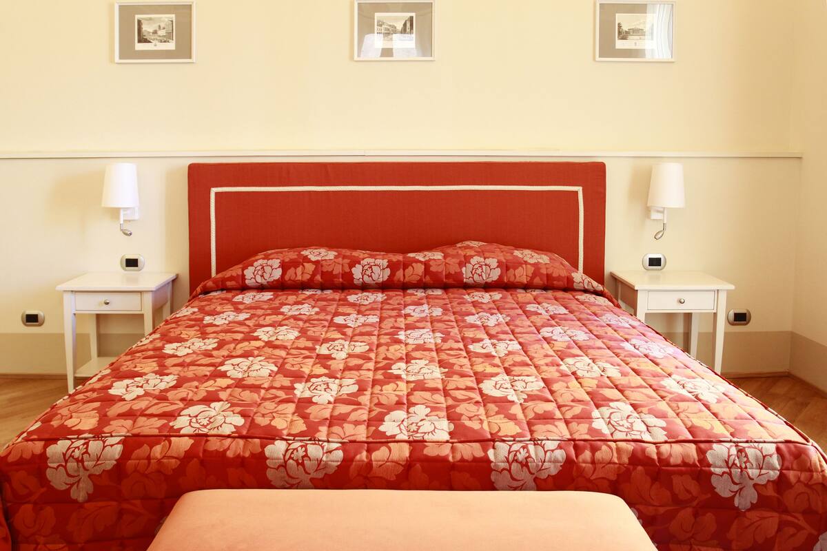 Un dormitorio ordenado con colcha de estampado floral, dos mesitas de noche con lámparas y cuadros en la pared.