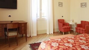 A hotel room with a wooden desk, two red armchairs, a bed with a floral bedspread, a small table with a lamp, and a TV mounted on the wall. - Casa Isolani Piazza Maggiore (Bologna)