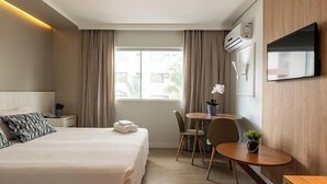 Habitación de hotel con cama, escritorio, silla, una mesita con una planta, televisor montado en la pared y ventana con cortinas.