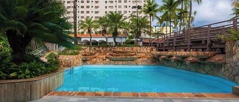 A swimming pool with a stone wall and palm trees, surrounded by a paved area.