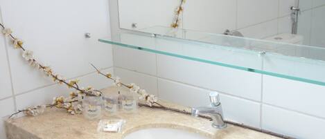 Un baño con un lavamanos blanco, un estante de vidrio sobre el lavamanos y un espejo que refleja una rama con flores.