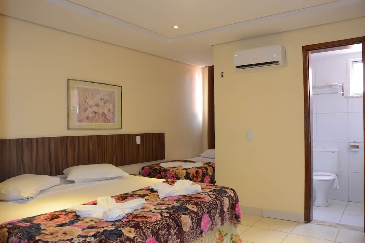 Ein Hotelzimmer mit zwei Betten, einem hölzernen Kopfteil, einem gerahmten Blumenbild, einer Klimaanlage und einem Badezimmer mit Toilette und Handtuchhalter.