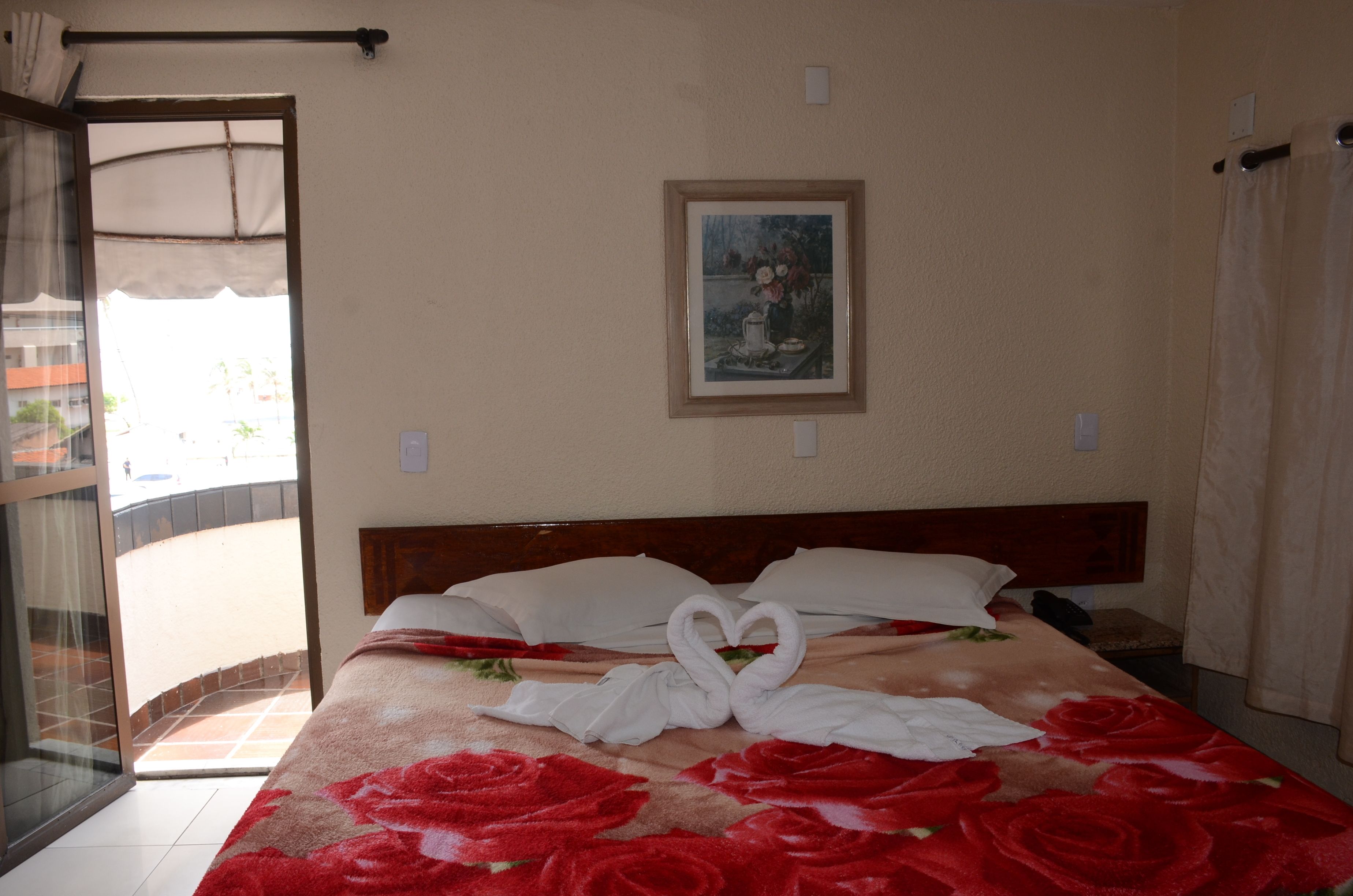 Habitación de hotel con cama, colcha estampada de flores rojas, toallas dispuestas en forma de corazón y un cuadro en la pared.
