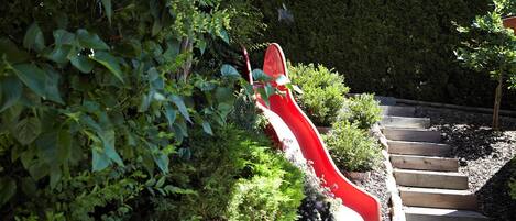 Un tobogán rojo en un jardín, con una escalera de madera que sube hasta él.
