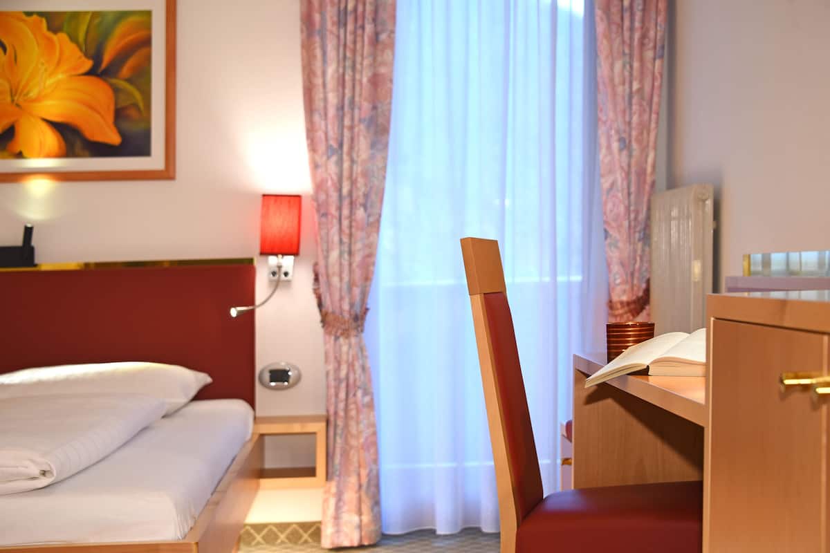 A hotel room with a bed, a desk, a chair, a lamp, a painting, and a window with curtains.