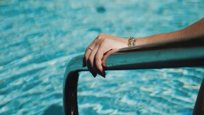 A hand resting on a poolside railing with clear blue water and a rocky shoreline in the background. - Hotel Hubertushof (Naz Sciaves)