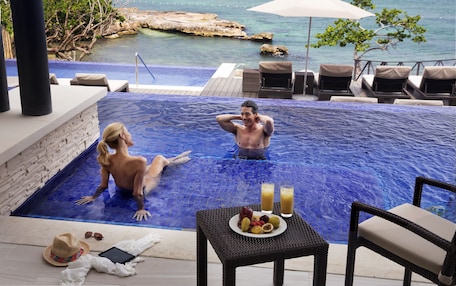 A couple relaxing in a private infinity pool with a view of the ocean, a table with fruit and drinks, and lounge chairs.