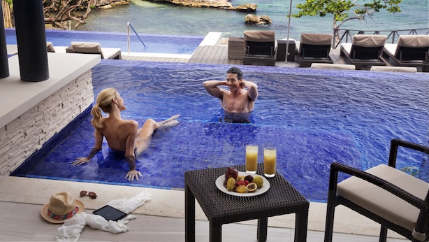 Een koppel ontspant in een privé-overloopzwembad met uitzicht op de oceaan, een tafel met fruit en drankjes, en ligstoelen.