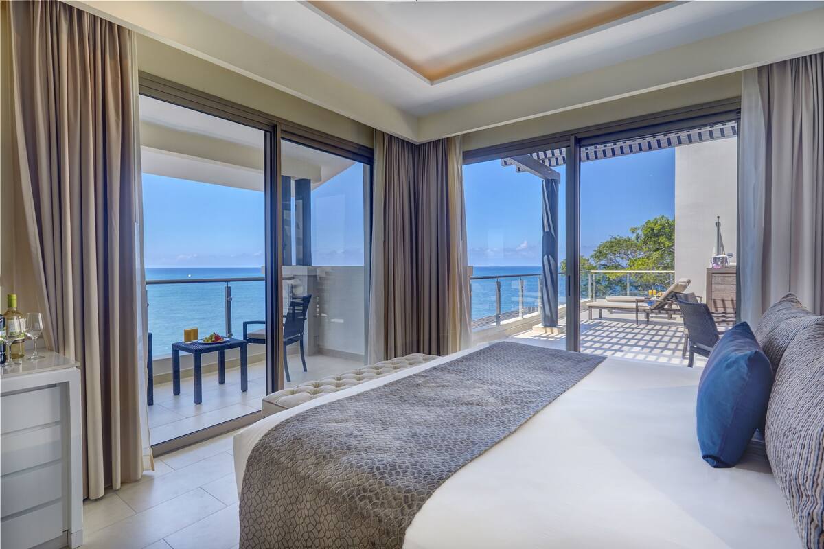Une chambre d’hôtel dotée d’un grand lit, d’un balcon avec vue sur la mer et d’un coin repas avec des sièges en plein air.