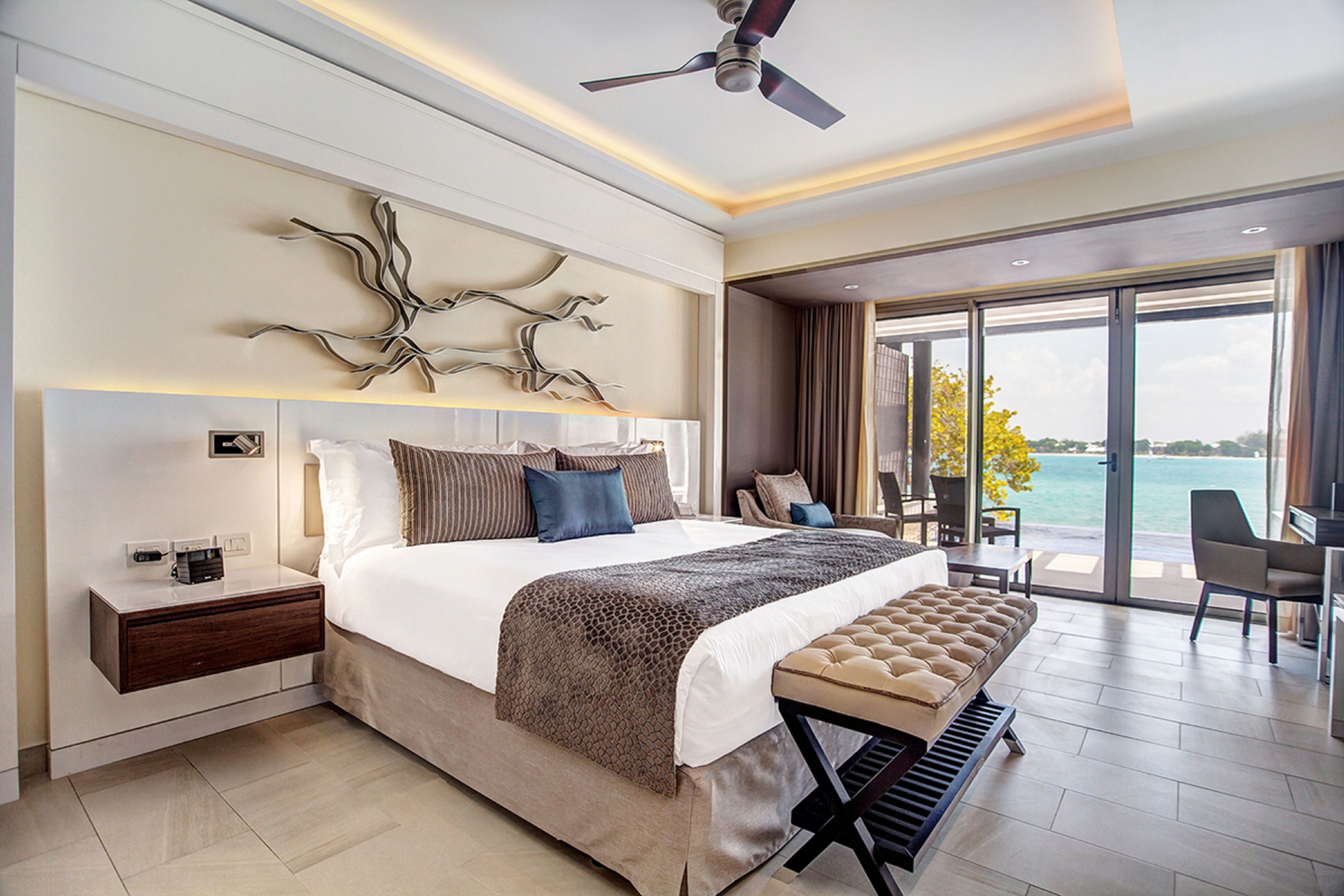 A modern hotel room with a large bed, a bench, a desk, and a view of the sea.