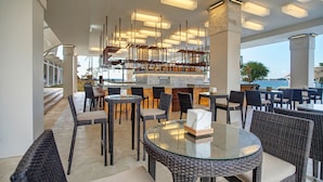 Een terras met een bar, diverse zithoeken zoals ronde tafels en rieten stoelen, en een bar met een uitgebreid assortiment flessen.
